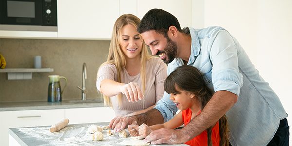 Mom and dad teaching daughter to make dough on kitchen table with flour